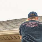 Man on a ladder at a roofline, seen from behind, wearing a dark T-shirt that reads 'Always on Top' with a Roof Raiders logo.
