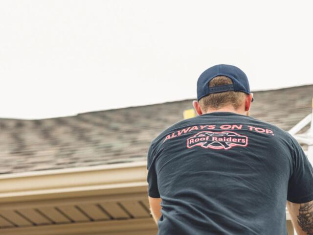 Man on a ladder at a roofline, seen from behind, wearing a dark T-shirt that reads 'Always on Top' with a Roof Raiders logo.