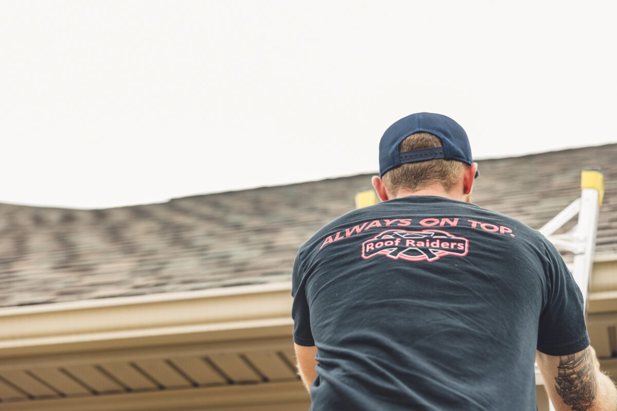 Man on a ladder at a roofline, seen from behind, wearing a dark T-shirt that reads 'Always on Top' with a Roof Raiders logo.