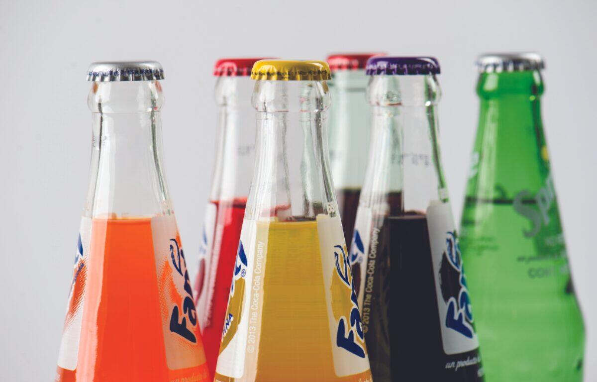 Close-up of several glass soda bottles with colorful liquids (orange, red, yellow, dark, and green) lined up in a row.