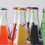 Close-up of several glass soda bottles with colorful liquids (orange, red, yellow, dark, and green) lined up in a row.