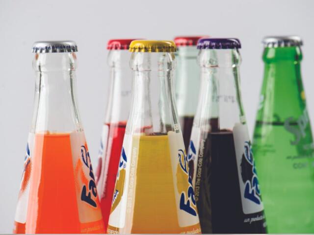 Close-up of several glass soda bottles with colorful liquids (orange, red, yellow, dark, and green) lined up in a row.