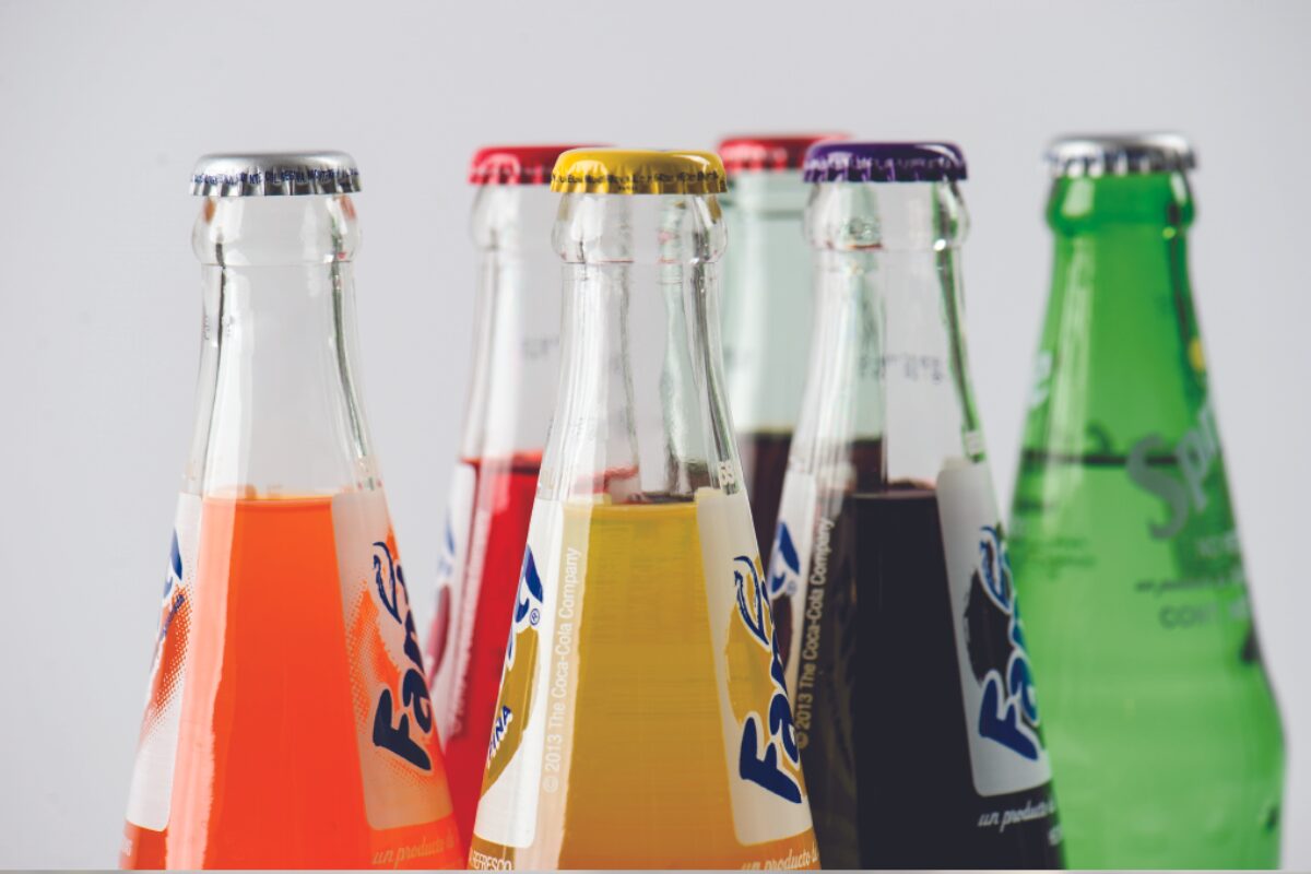 Close-up of several glass soda bottles with colorful liquids (orange, red, yellow, dark, and green) lined up in a row.