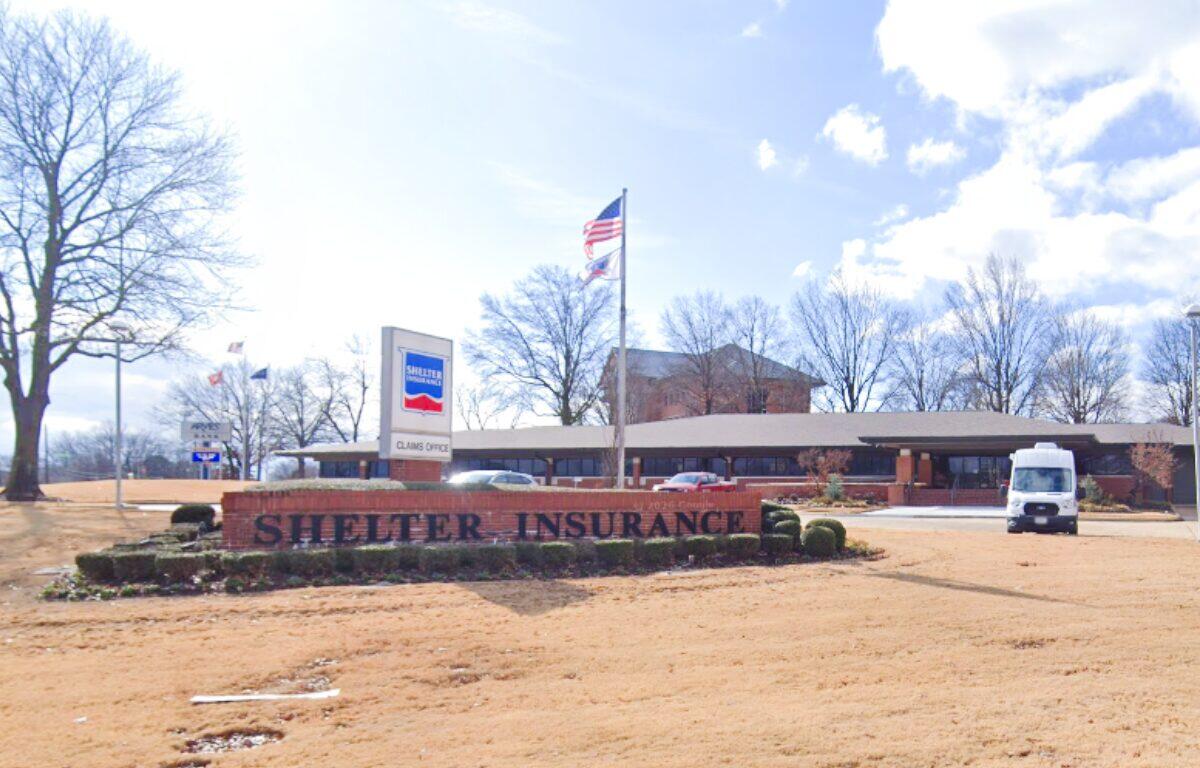 Shelter Insurance office with a brick sign in front reading 'Shelter Insurance' and an American flag nearby; a white van is parked to the right.
