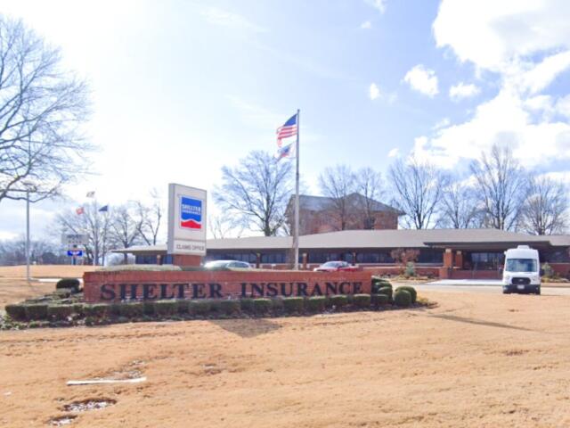 Shelter Insurance office with a brick sign in front reading 'Shelter Insurance' and an American flag nearby; a white van is parked to the right.