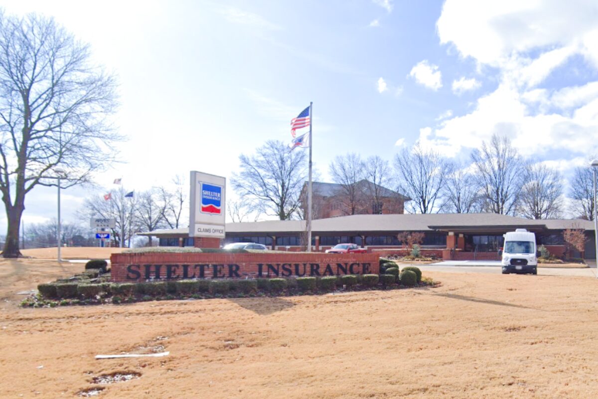 Shelter Insurance office with a brick sign in front reading 'Shelter Insurance' and an American flag nearby; a white van is parked to the right.