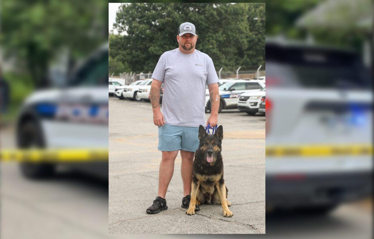 Man in a gray t-shirt and blue shorts stands next to a German Shepherd in a parking lot, both looking at the camera.