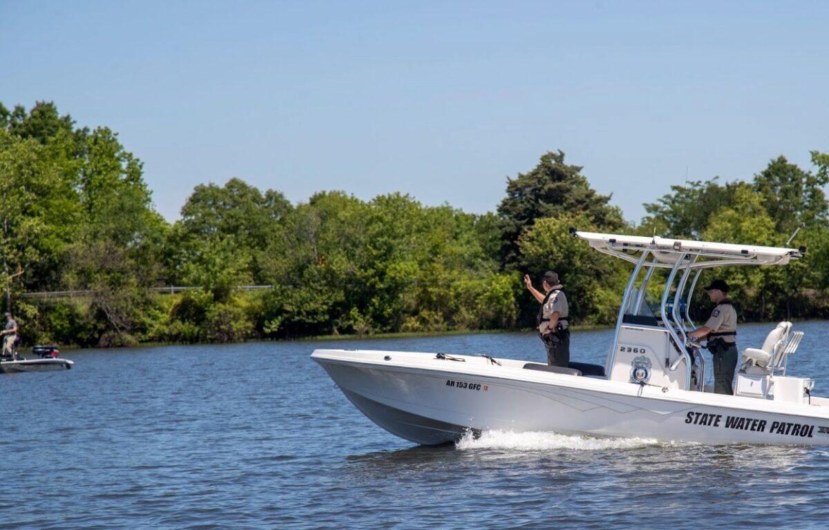 White state water patrol boat speeding on a calm lake, two officers on board with shoreline trees in the background.