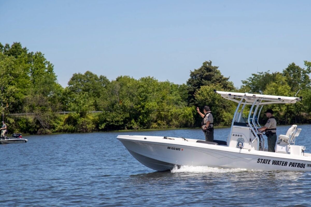 White state water patrol boat speeding on a calm lake, two officers on board with shoreline trees in the background.