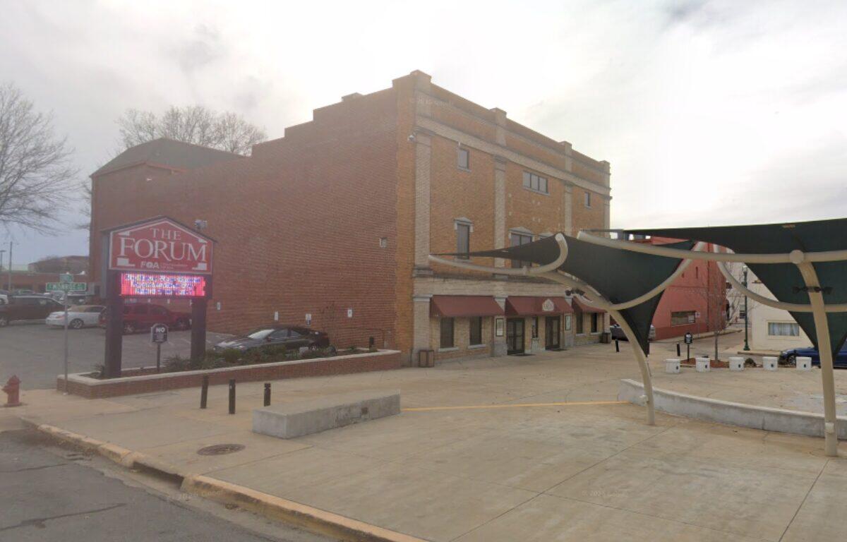 Brick building with storefronts and a large 'The Forum' sign, plus a digital marquee in front and a concrete plaza area.