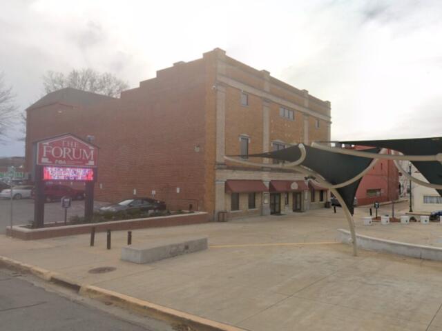 Brick building with storefronts and a large 'The Forum' sign, plus a digital marquee in front and a concrete plaza area.
