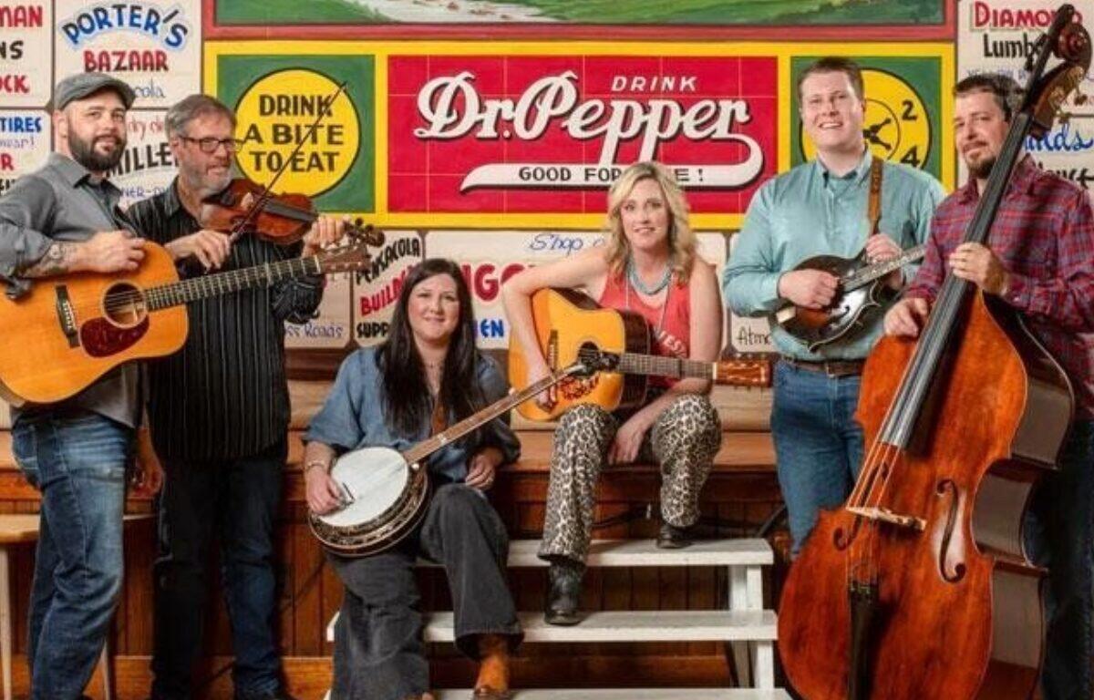 Six musicians with guitars, a fiddle, banjo and a bass pose in front of a colorful Dr Pepper mural on a stage bench scene in a retro setting.