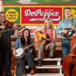 Six musicians with guitars, a fiddle, banjo and a bass pose in front of a colorful Dr Pepper mural on a stage bench scene in a retro setting.