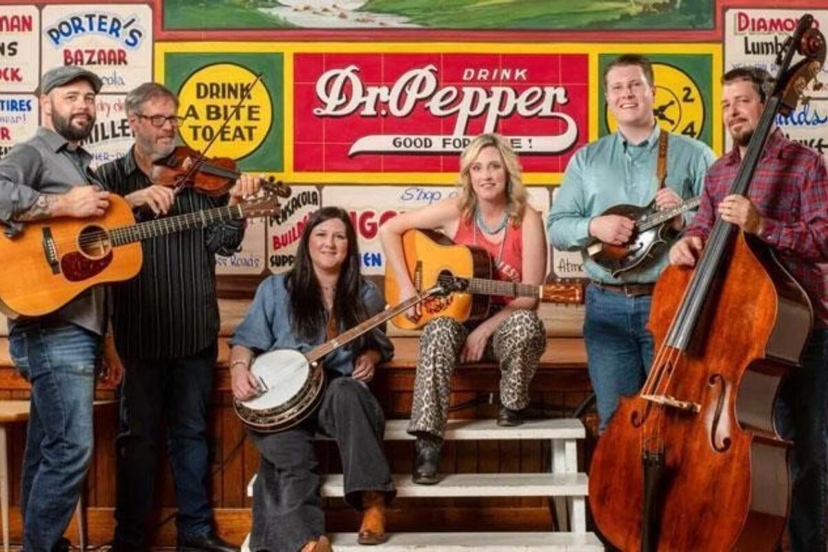Six musicians with guitars, a fiddle, banjo and a bass pose in front of a colorful Dr Pepper mural on a stage bench scene in a retro setting.
