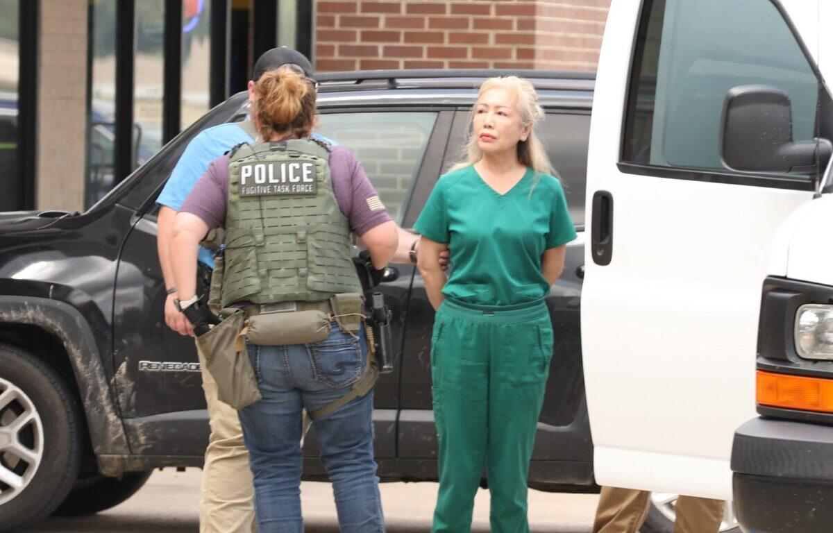 Police officer in tactical vest appears to detain a woman in green scrubs beside a black SUV outside a building.