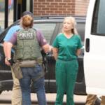 Police officer in tactical vest appears to detain a woman in green scrubs beside a black SUV outside a building.