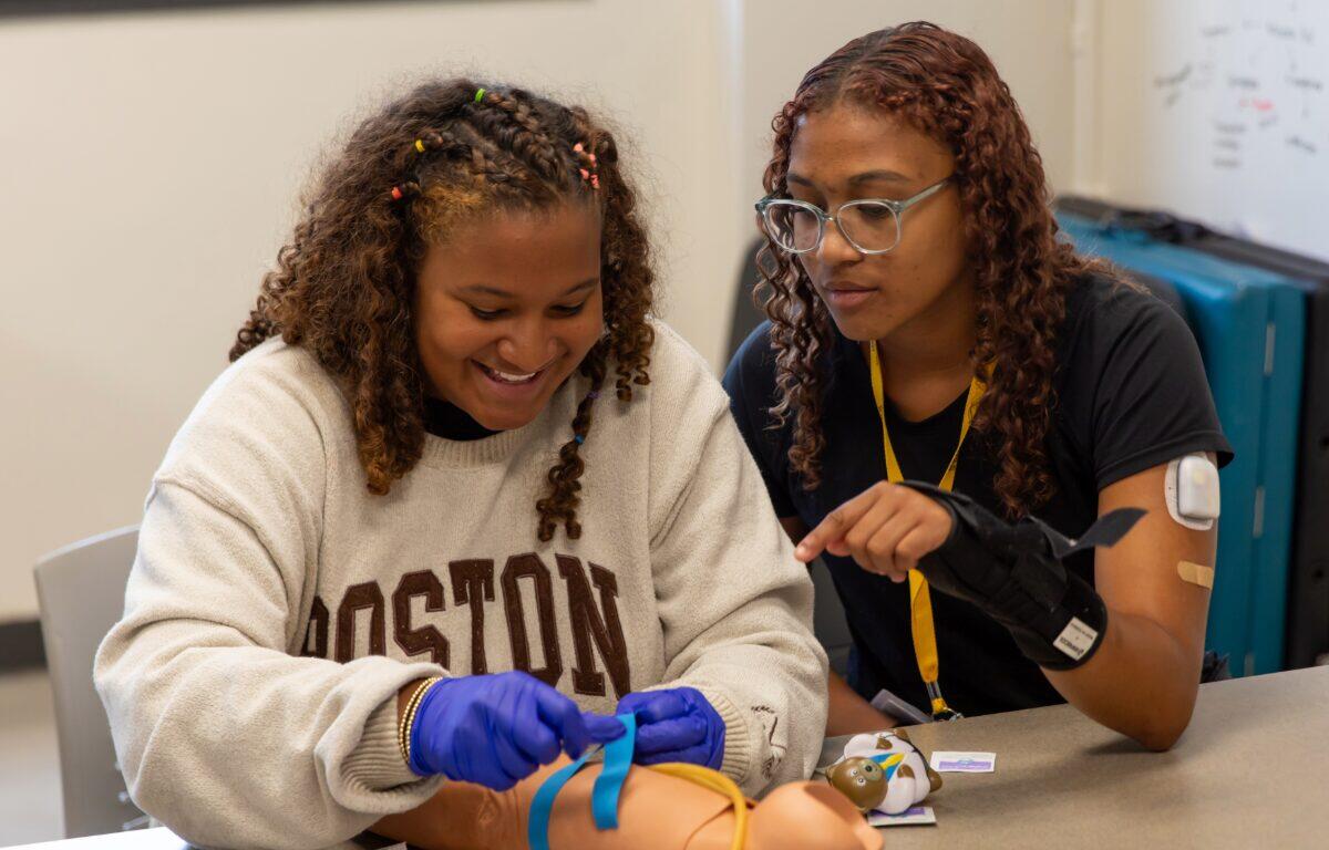 Two students practice medical training with a pediatric mannequin on a classroom table; one wears blue gloves and ties a strap while the other watches.