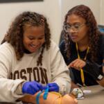 Two students practice medical training with a pediatric mannequin on a classroom table; one wears blue gloves and ties a strap while the other watches.