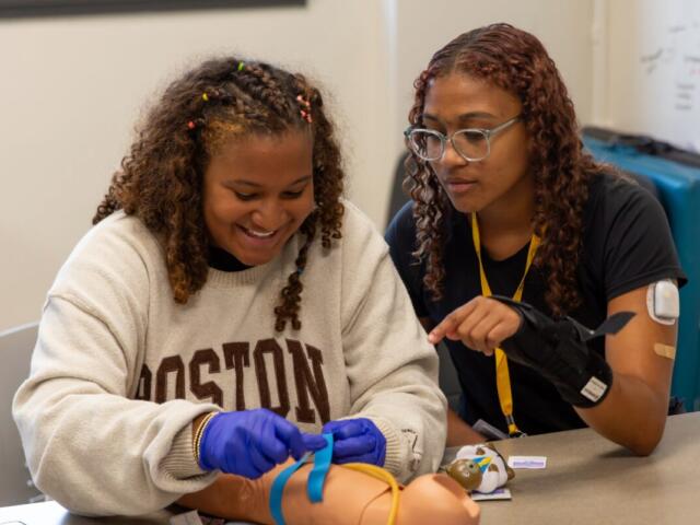Two students practice medical training with a pediatric mannequin on a classroom table; one wears blue gloves and ties a strap while the other watches.