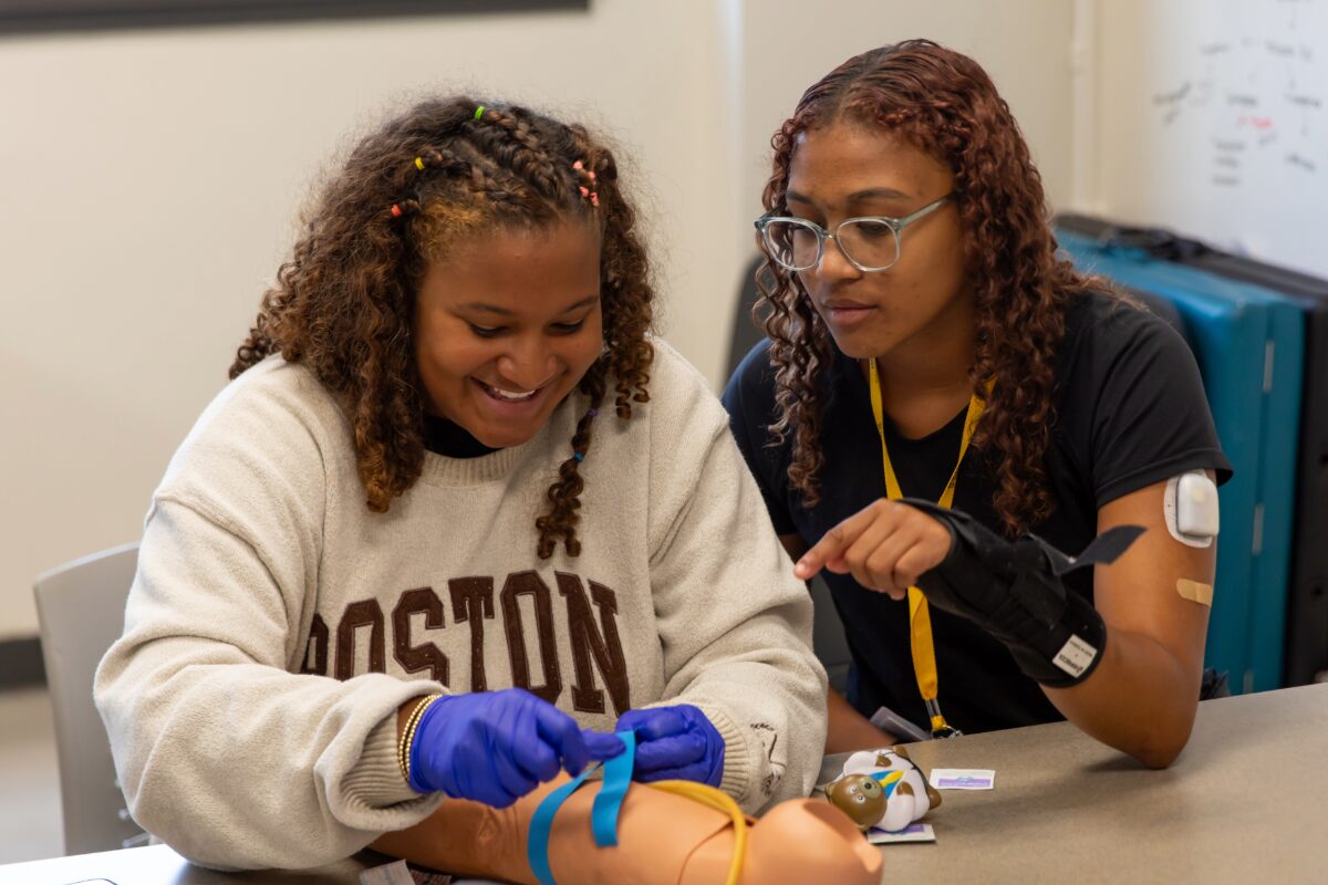 Two students practice medical training with a pediatric mannequin on a classroom table; one wears blue gloves and ties a strap while the other watches.