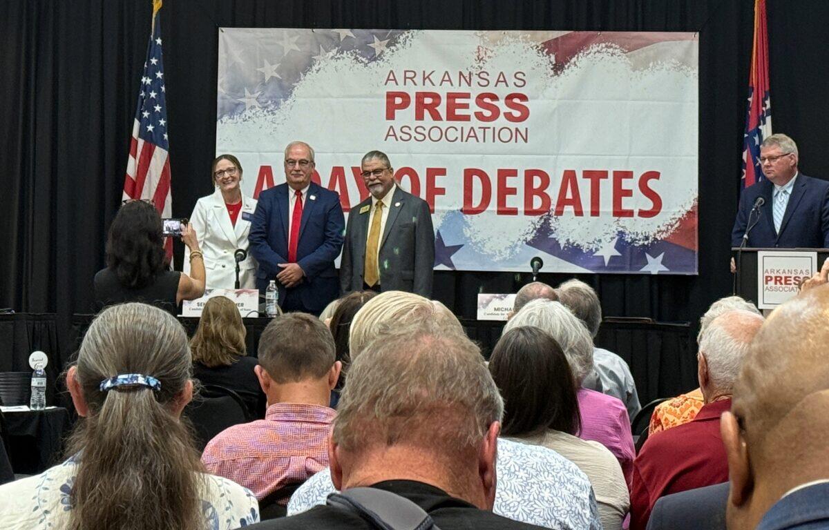 Panelists stand on a stage in front of a banner that reads Arkansas Press Association, during a debate event, with audience watching.