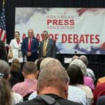 Panelists stand on a stage in front of a banner that reads Arkansas Press Association, during a debate event, with audience watching.