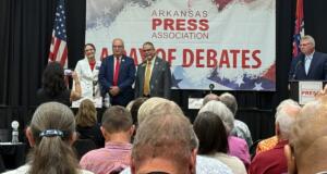 Panelists stand on a stage in front of a banner that reads Arkansas Press Association, during a debate event, with audience watching.