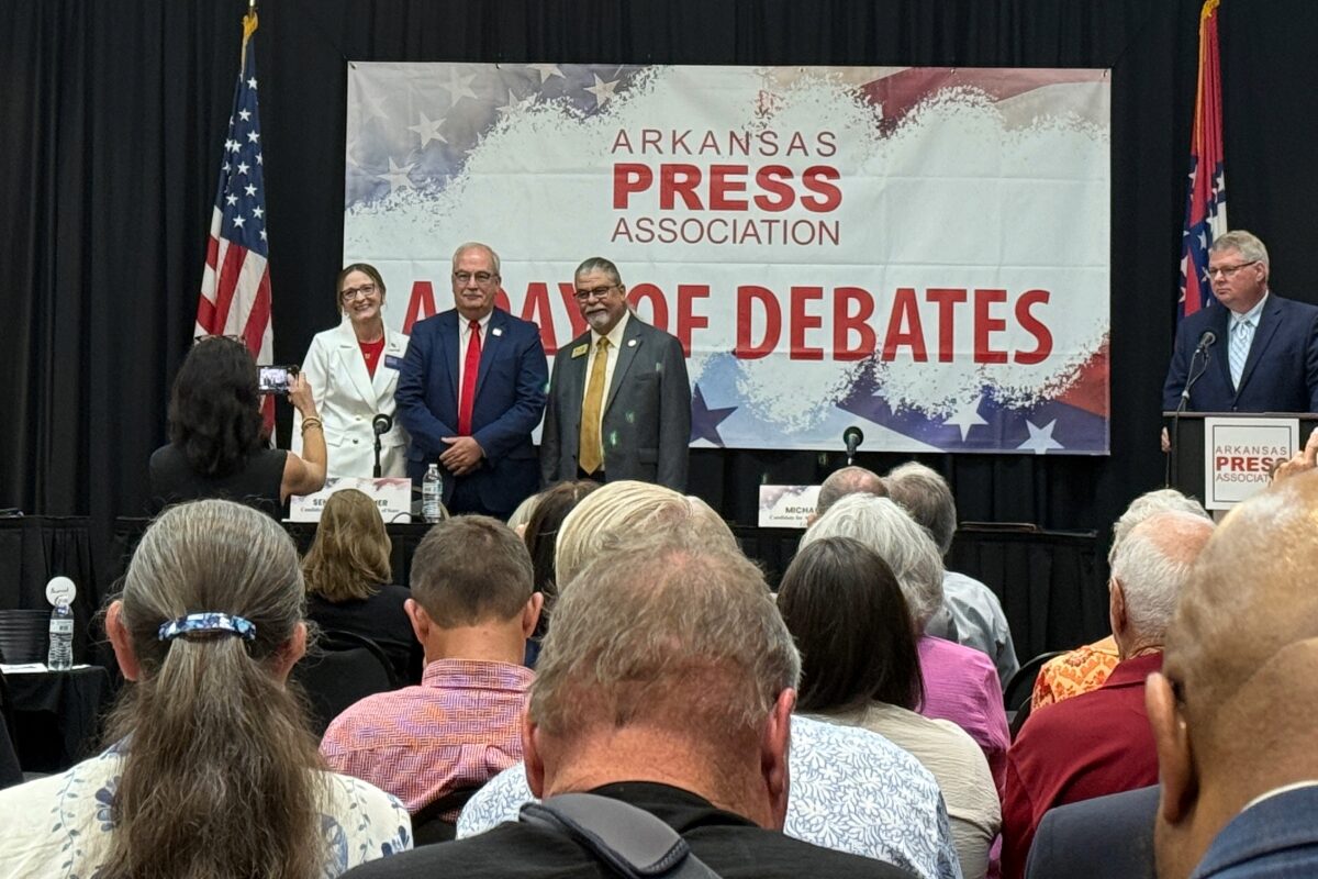 Panelists stand on a stage in front of a banner that reads Arkansas Press Association, during a debate event, with audience watching.