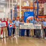 Group of people in a warehouse holding 'Million Meals' signs and a large donation check, highlighting a hunger relief event.