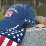 Blue veteran cap with a white 'VETERAN' on the brim and a US flag patch rests on a granite tombstone, beside a star-spangled scarf in a cemetery.