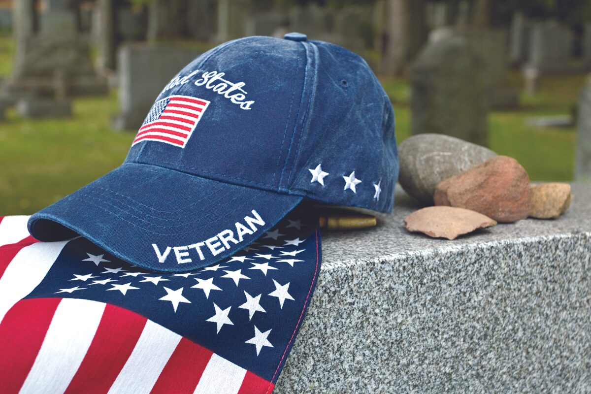 Blue veteran cap with a white 'VETERAN' on the brim and a US flag patch rests on a granite tombstone, beside a star-spangled scarf in a cemetery.