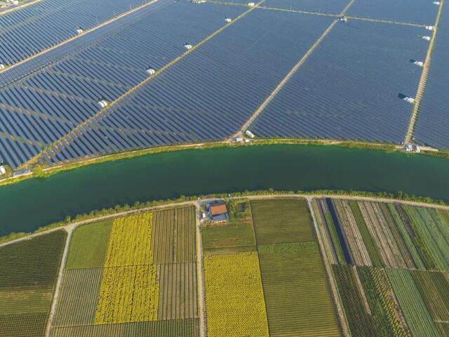 Aerial view of a large solar farm separated from colorful patchwork fields by a winding canal.