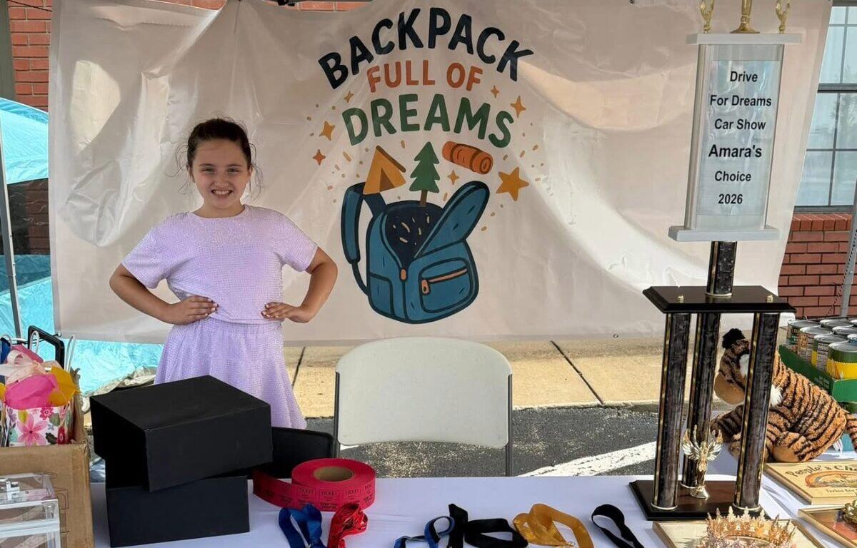 Young girl in a lavender dress posing with hands on hips at a charity booth that reads 'Backpack Full of Dreams' with a blue backpack illustration on a backdrop.