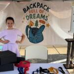 Young girl in a lavender dress posing with hands on hips at a charity booth that reads 'Backpack Full of Dreams' with a blue backpack illustration on a backdrop.