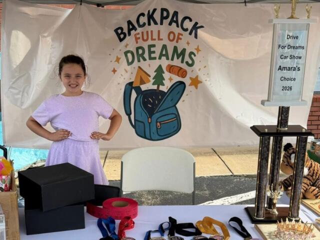 Young girl in a lavender dress posing with hands on hips at a charity booth that reads 'Backpack Full of Dreams' with a blue backpack illustration on a backdrop.