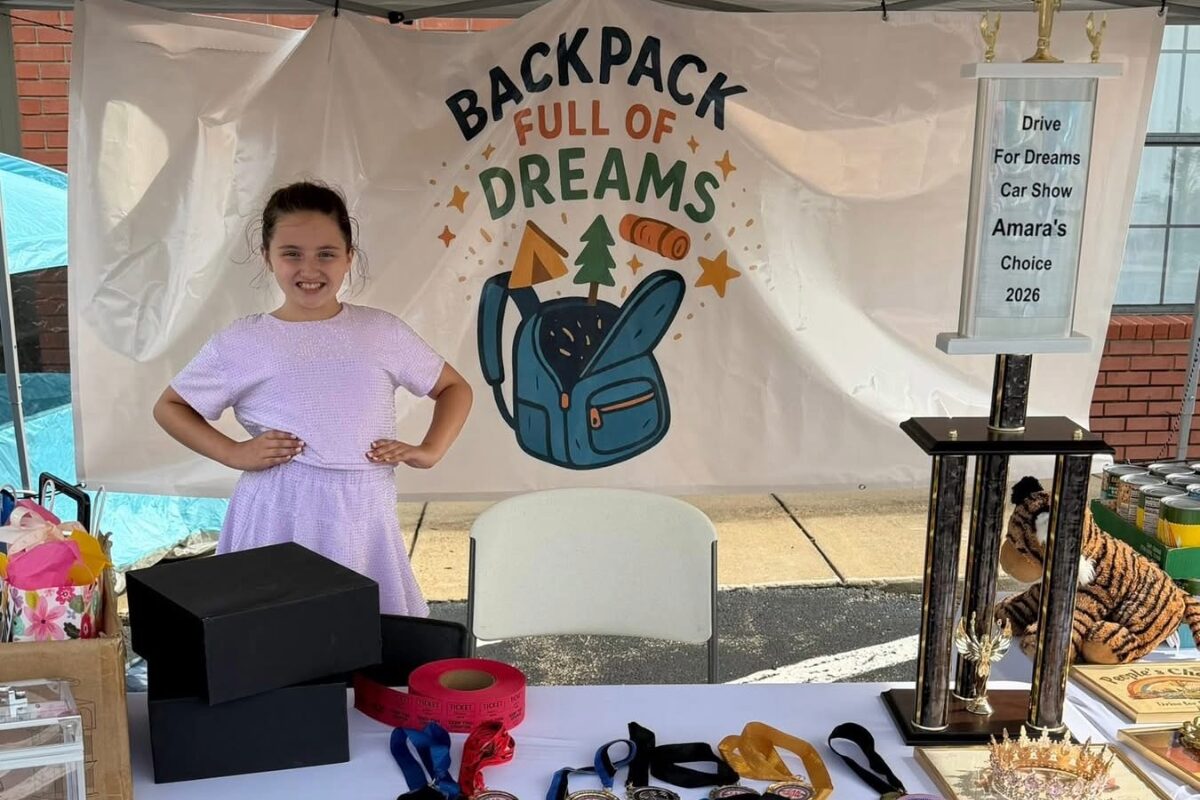 Young girl in a lavender dress posing with hands on hips at a charity booth that reads 'Backpack Full of Dreams' with a blue backpack illustration on a backdrop.