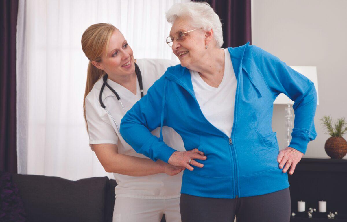 Caregiver helps a smiling elderly woman stand, placing one hand on her lower back in a living room setting.