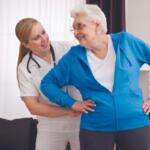 Caregiver helps a smiling elderly woman stand, placing one hand on her lower back in a living room setting.
