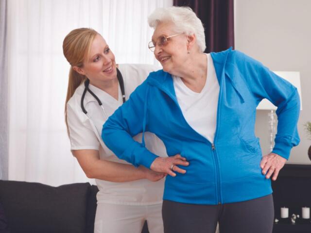 Caregiver helps a smiling elderly woman stand, placing one hand on her lower back in a living room setting.