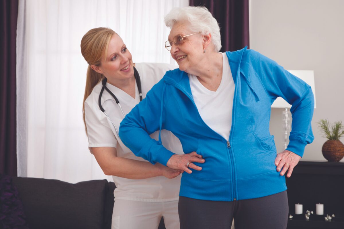 Caregiver helps a smiling elderly woman stand, placing one hand on her lower back in a living room setting.