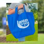 Blue tote bag with 'Library 2 Go' logo being carried outdoors. I