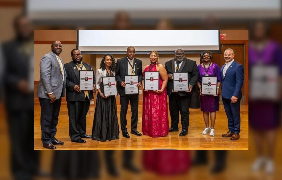 Group of eight people in formal attire posing on a stage, each holding a framed certificate or award.