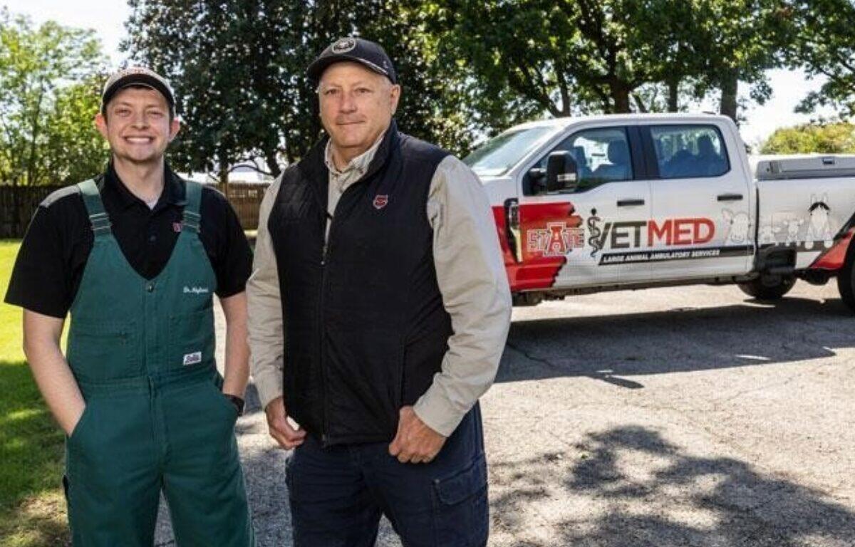 Two men posing outdoors beside a white pickup with an ETMED emergency services logo, one in green overalls and the other in a black vest and cap.
