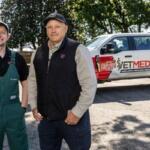 Two men posing outdoors beside a white pickup with an ETMED emergency services logo, one in green overalls and the other in a black vest and cap.