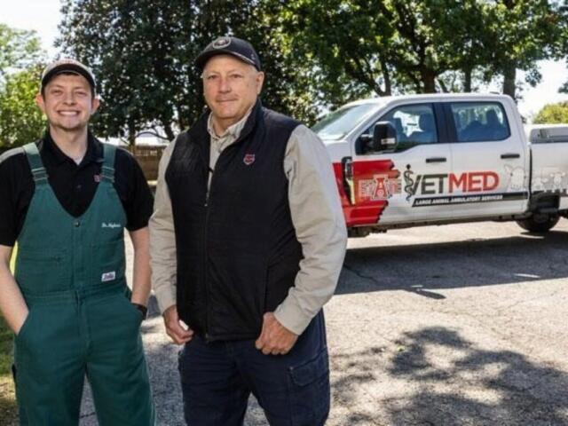 Two men posing outdoors beside a white pickup with an ETMED emergency services logo, one in green overalls and the other in a black vest and cap.