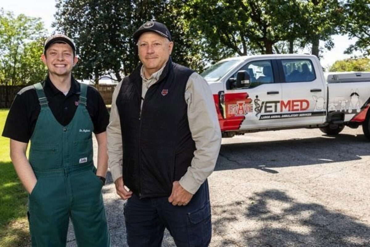 Two men posing outdoors beside a white pickup with an ETMED emergency services logo, one in green overalls and the other in a black vest and cap.