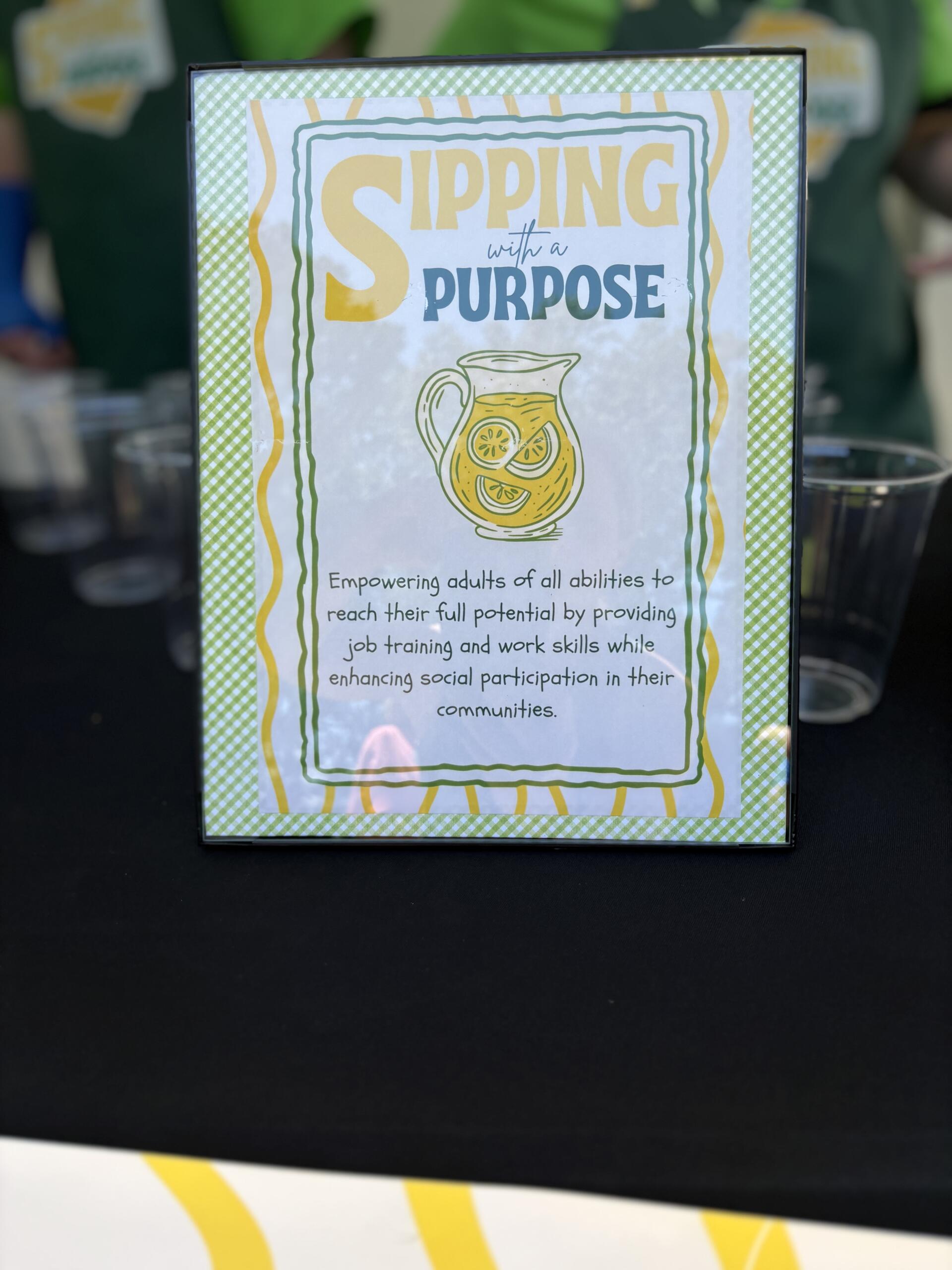 Poster reading 'Sipping with a Purpose' with a lemonade pitcher illustration and a green checkered border framing the sign