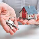 Hands cupping a set of keys beside a small red-and-white house model, symbolizing home purchase or real estate access