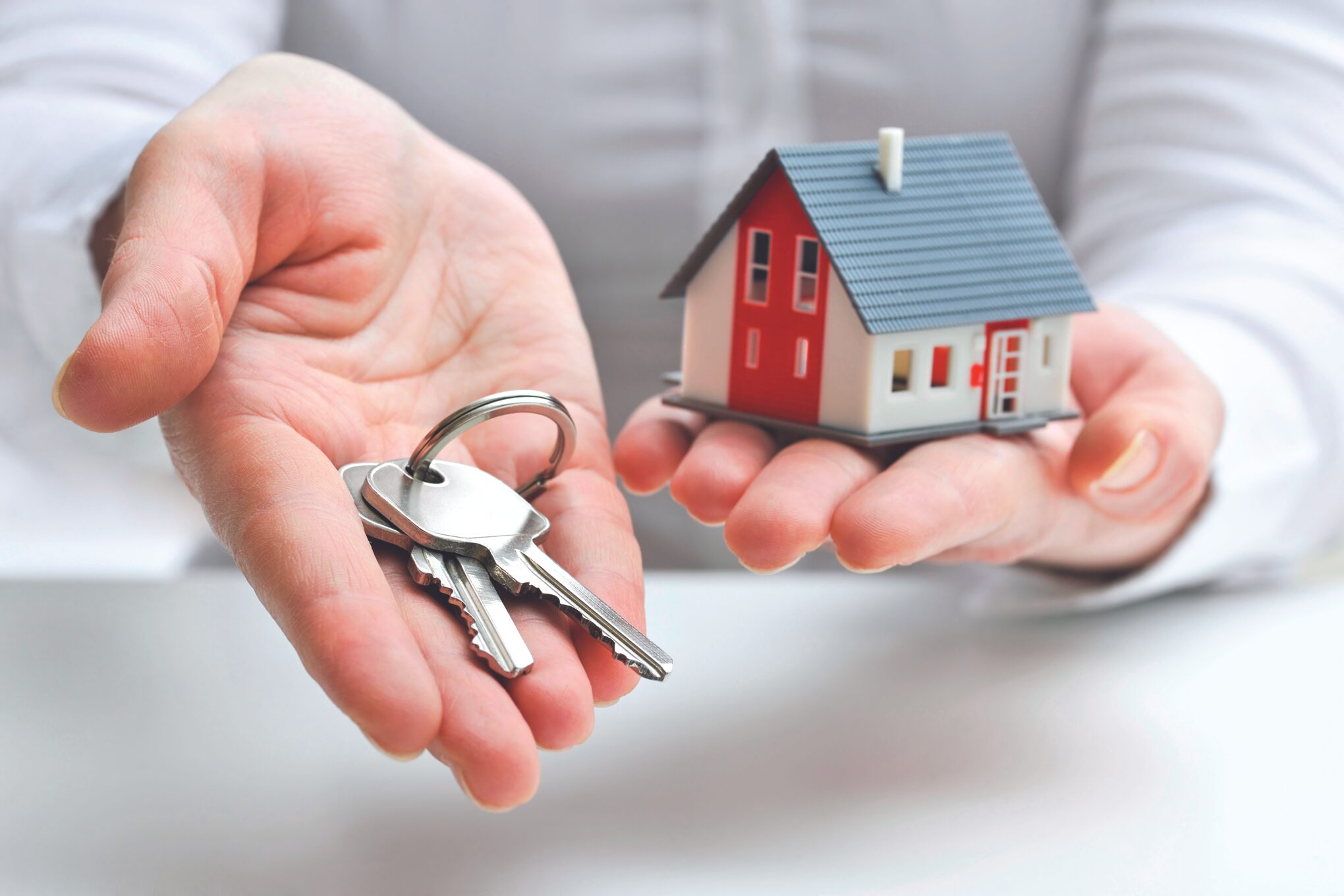 Hands cupping a set of keys beside a small red-and-white house model, symbolizing home purchase or real estate access