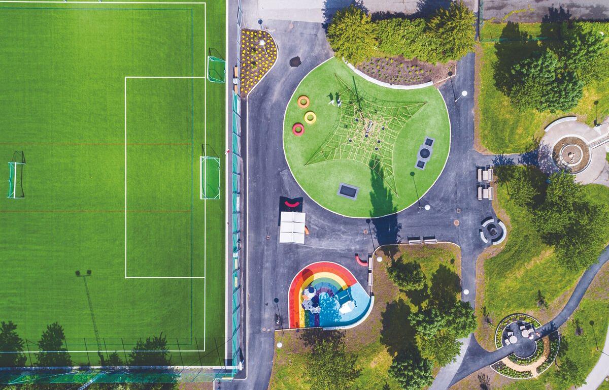 Aerial view of a sports field on the left and a park playground on the right, with a climbing net, rainbow slide, and green, circular play area.